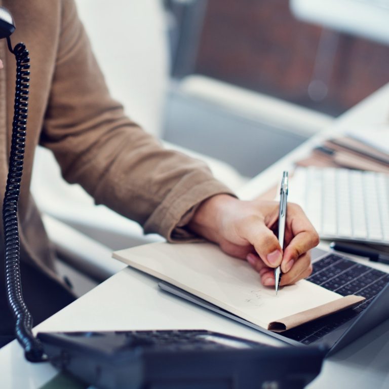 Vorteile für eine Messie Entrümpelung Berlin Eine Hand schreibt auf einem Notizblock, während ein Laptop und Telefon sichtbar sind.